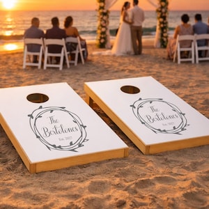Könnte beinhalten: Zwei weiße Cornhole-Bretter aus Holz mit naturfarbenen Kanten, jeweils mit einem kreisförmigen Loch und einem dunkelgrauen Kranz-Grafikdesign, das den Text "The Bertolones Est. 2027" auf der Oberfläche umschließt, platziert auf einem Sandstrand.