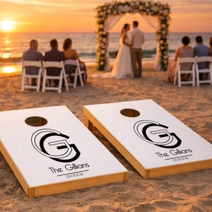 Könnte beinhalten: Zwei weiße Cornhole-Boards mit Seiten aus Naturholz, jeweils mit einem schwarzen kreisförmigen Monogramm mit dem Buchstaben G, dem Text The Gillians und dem Datum 05.05.2026 auf der Oberfläche, platziert auf einem Sandstrand bei Sonnenuntergang.