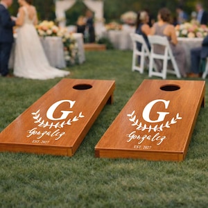 Può includere: Una coppia di tavole da cornhole in legno con finitura marrone naturale e un foro circolare nella parte superiore. Ogni tavola presenta una grafica bianca con una grande lettera G, un disegno a corona d'alloro, il nome Gonzalez e EST. 2027.