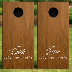 Könnte beinhalten: Zwei rechteckige Cornhole-Spielbretter aus Holz mit dunkelbrauner Holzmaserung. Jedes Brett hat oben ein kreisförmiges Loch. Das linke Brett zeigt den weißen Schriftzug "TEAM Bride" und das rechte "TEAM Groom" mit geschwungenen Verzierungen.