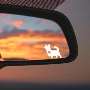 May include: A white Corgi dog decal with a heart detail, placed on a car's rearview mirror. The background shows a sunset with orange, yellow, and blue colours.