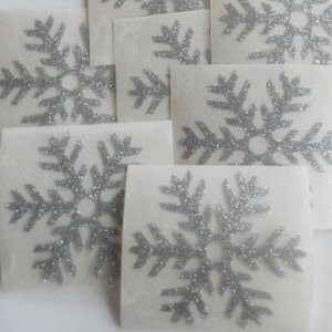 Puede incluir: Un conjunto de ocho pegatinas de copo de nieve de purpurina plateada sobre cartulina blanca.