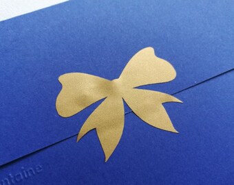 Può includere: Una busta blu reale con un design a fiocco dorato. Il fiocco è centrato sul lembo della busta. La busta è realizzata in carta strutturata. Il nome del marchio è impresso nell'angolo in basso a sinistra.