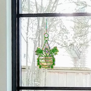 May include: A green and gold hanging planter with a geometric design is suspended in front of a window. The planter is filled with greenery and is hanging from a thin, light green string. The window has a black frame.