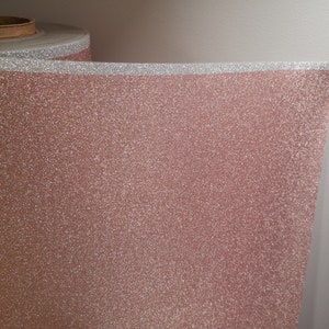 Può includere: Un rotolo di carta glitterata color oro rosa con un bordo glitterato argentato. La carta ha una superficie strutturata ed è parzialmente srotolata, rivelando il materiale glitterato. Lo sfondo è un muro bianco.