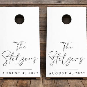 May include: Two white cornhole boards featuring a circular hole at the top. The boards are printed with the black text "The Stelzers" in a cursive script font, followed by the date "AUGUST 4, 2027" in a clean, sans-serif capital font.