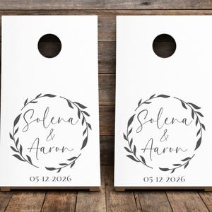 Könnte beinhalten: Zwei weiße Cornhole-Boards mit einem kreisförmigen, dunkelgrauen Blattkranz-Design. Innerhalb des Kranzes sind die Namen Solena & Aaron in Schreibschrift gedruckt. Unter dem Kranz steht das Datum 05-12-2026 in einer Serifenschrift.