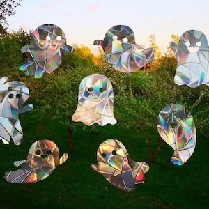 May include: Nine iridescent holographic ghost cutouts with smiling faces are arranged in a circle on a green grassy background with trees in the background.