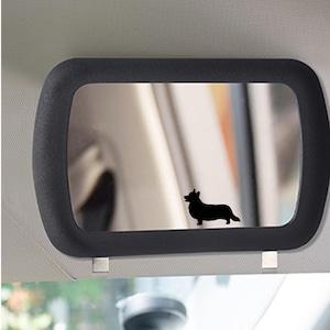 May include: A rectangular car visor mirror featuring a thick black frame and a reflective surface. A solid black silhouette of a Corgi dog is positioned on the lower right side of the mirror glass, adding a distinct graphic element to the design.
