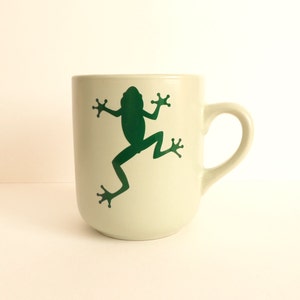 May include: A cream-colored ceramic mug with a green frog silhouette design. The frog is depicted in a leaping pose. The mug has a curved handle and a simple, cylindrical shape, suitable for hot or cold beverages.