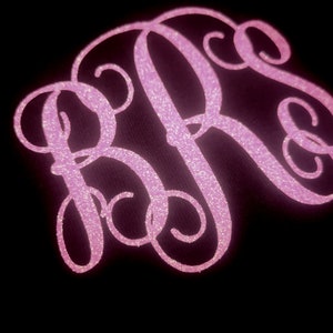 Może przedstawiać: Różowy brokatowy monogram z przeplatającymi się literami R i S na czarnym tle. Litery mają kursywny, elegancki styl, z błyszczącym, teksturowanym wyglądem. Ogólny projekt jest dekoracyjny i przyciągający wzrok.