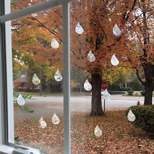 Puede incluir: Una ventana con vista a un árbol con follaje otoñal. La ventana está decorada con pegatinas de gotas de lluvia arcoíris iridiscentes. Las pegatinas están dispuestas en un patrón aleatorio en la ventana. La palabra "STOP" es visible en un letrero en el fondo.