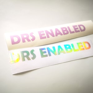 May include: Two rectangular stickers with the text "DRS ENABLED". One sticker has light purple text on a cream background. The other sticker has iridescent text on a white background. The stickers are likely for a car or other vehicle.