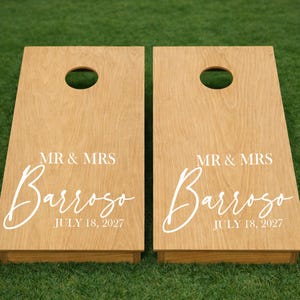 May include: Two light brown wooden cornhole boards featuring a circular hole at the top. Each board displays the white printed text MR & MRS, the name Barroso in a large script font, and the date JULY 18, 2027, centred on the surface.