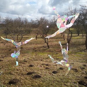 Puede incluir: Tres figuras iridiscentes se recortan sobre un fondo de árboles desnudos y un cielo nublado. Las figuras están en varias poses, lo que sugiere una danza o una rutina de gimnasia. Las figuras están hechas de un material reflectante que crea un efecto brillante.