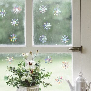 May include: A window with white trim and a white teapot on a windowsill. The window is decorated with iridescent snowflake stickers. A bouquet of white flowers sits in a white vase on the windowsill.