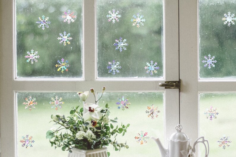 May include: A window with white trim and a white teapot on a windowsill. The window is decorated with iridescent snowflake stickers. A bouquet of white flowers sits in a white vase on the windowsill.