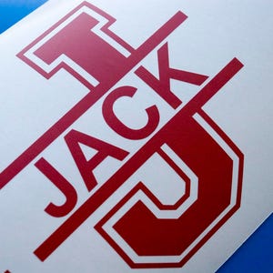 Może przedstawiać: Czerwona naklejka winylowa z napisem "JACK" napisanym pogrubioną czcionką z dużą czerwoną literą "J" w stylu uniwersyteckim.