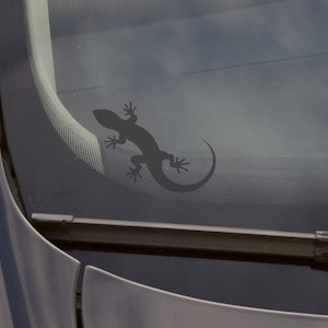 Pode incluir: Uma silhueta preta de um lagarto está no pára-brisas de um carro. O lagarto está olhando para o lado direito da imagem.