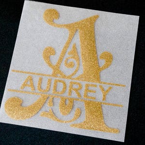 Könnte beinhalten: Goldener Glitzer-Vinyl-Aufkleber mit einem dekorativen Buchstaben A und dem Namen "Audrey" in einem Banner darunter.