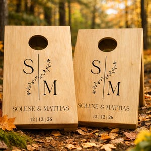 Könnte beinhalten: Zwei helle Holz-Cornhole-Boards mit einem kreisförmigen Loch an der Oberseite. Jedes Board ist mit den schwarzen Buchstaben S und M, getrennt durch eine vertikale Rankengrafik, den Namen SOLENE & MATTIAS und dem Datum 12 | 12 | 26 bedruckt.