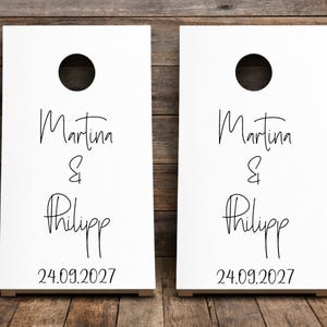 Könnte beinhalten: Zwei weiße Cornhole-Boards mit einem kreisförmigen Loch im oberen Bereich. Die Boards sind mit dem schwarzen Schriftzug Martina & Philipp in einer Schreibschrift bedruckt, gefolgt von dem Datum 24.09.2027 in einer serifenlosen Schrift.