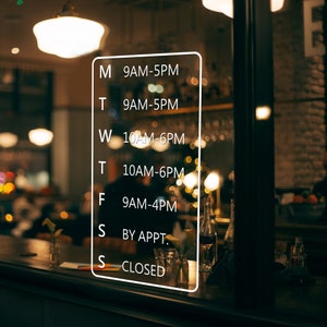 以下が含まれることがあります： 週ごとの営業時間を示す、黒い文字で書かれた白い看板。看板には、月曜日午前9時～午後5時、火曜日午前9時～午後5時、水曜日午前10時～午後6時、木曜日午前10時～午後6時、金曜日午前9時～午後4時、土曜日予約制、日曜日休業と記載されている。