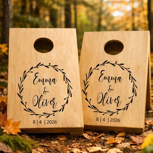 Könnte beinhalten: Zwei helle Holz-Cornhole-Boards mit einem kreisförmigen Loch an der Oberseite. Jedes Board zeigt einen schwarzen, aufgedruckten Kranz um die Namen Emma & Oliver, mit dem Datum 8 | 4 | 2026 in Schwarz unter dem Kranz.