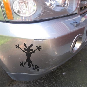 May include: A silver car with a black silhouette of a coyote sticker on the front bumper. The sticker is of a coyote with its arms raised in the air.
