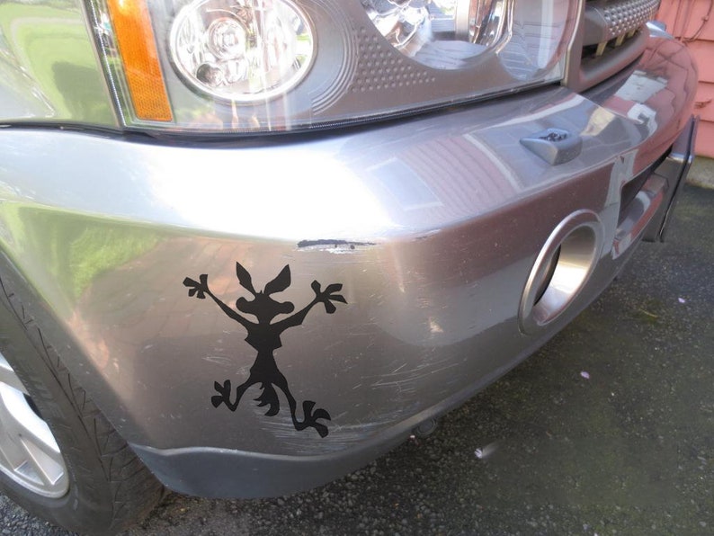 May include: A silver car with a black silhouette of a coyote sticker on the front bumper. The sticker is of a coyote with its arms raised in the air.