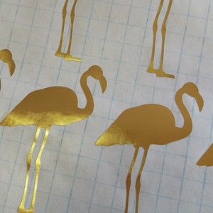 May include: Gold metallic flamingo silhouette stickers. The stickers are on a white gridded background.