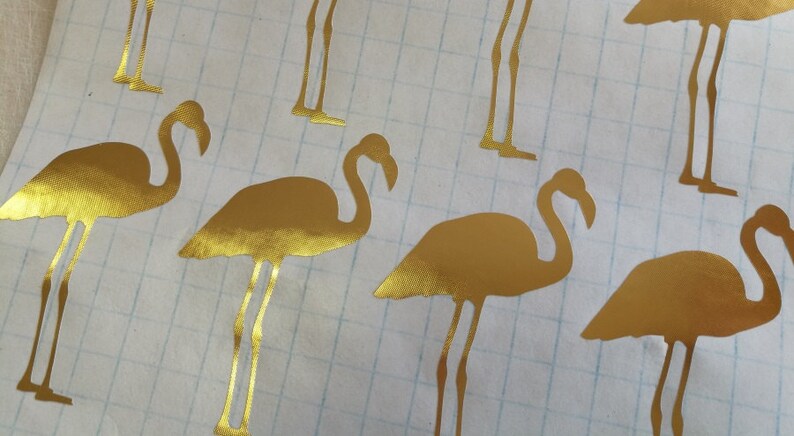 May include: Gold metallic flamingo silhouette stickers. The stickers are on a white gridded background.