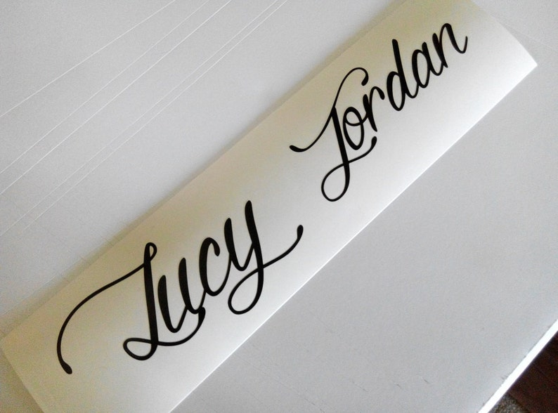 Op de afbeelding: Witte vinyl sticker met de namen "Lucy" en "Jordan" geschreven in zwarte cursieve letters.