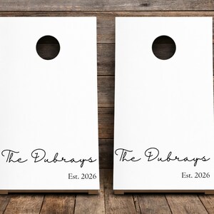 Könnte beinhalten: Zwei weiße Cornhole-Boards mit einem kreisförmigen Loch im oberen Bereich. Jedes Board zeigt den schwarzen kursiven Schriftzug The Dubrays und den gedruckten Text Est. 2026 in einer einfachen Serifenschrift unterhalb des Namens.