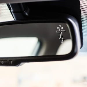 May include: A white Eastern Orthodox cross sticker is attached to a car's rearview mirror. The cross design has a top bar, a larger middle bar, and a slanted bottom bar. The mirror is black and reflects the car's interior.