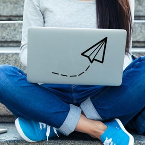 May include: A person sitting with a laptop computer on their lap. The laptop has a paper aeroplane sticker on the lid with a dotted line trailing behind it.