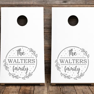 Könnte beinhalten: Zwei weiße Cornhole-Boards mit einer kreisförmigen grauen Grafik und einem Blattkranz-Design. Der Text im Kreis lautet the WALTERS family in einer Mischung aus Serif- und Schreibschrift, akzentuiert durch kleine Punkte. Jedes Board hat ein Loch.