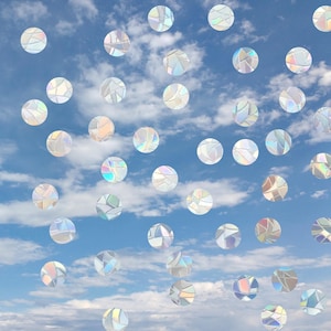 May include: A collection of circular window decals featuring a prismatic, iridescent finish that reflects light in various colors. The stickers are arranged in a scattered pattern against a backdrop of a bright blue sky with white clouds.