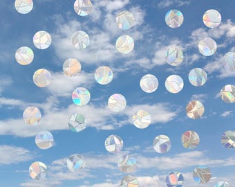 May include: A collection of circular window decals featuring a prismatic, iridescent finish that reflects light in various colors. The stickers are arranged in a scattered pattern against a backdrop of a bright blue sky with white clouds.
