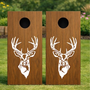 Könnte beinhalten: Zwei rechteckige Cornhole-Bretter mit einer dunkelbraunen Holzmaserung. Jedes Brett hat ein kreisförmiges Loch im oberen Bereich und eine große, weiße, stilisierte Grafik eines Hirschkopfes mit Geweih in der Mitte der vorderen Holzoberfläche.