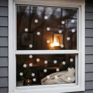 Puede incluir: Una ventana de doble guillotina con numerosos adhesivos de copos de nieve blancos aplicados sobre los cristales. La ventana tiene un marco blanco y está instalada en un revestimiento horizontal gris. Los adhesivos están dispersos.