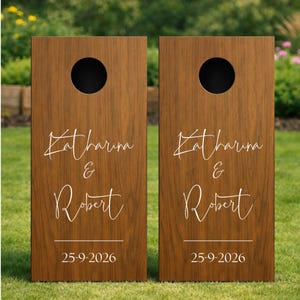 Könnte beinhalten: Zwei rechteckige Cornhole-Bretter aus Holz mit einer dunkelbraunen Holzmaserung. Jedes Brett hat oben ein kreisförmiges Loch und einen weißen Schriftzug Katharina & Robert mit dem Datum 25-9-2026 unter den Namen.