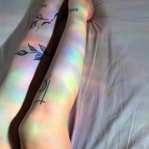 May include: A person's legs with tattoos are lying on a white bed sheet. The legs are illuminated by a rainbow prism light, creating a colourful effect.
