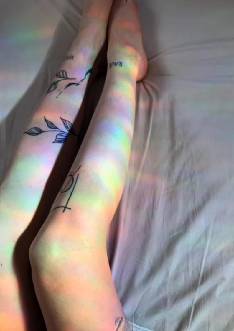 May include: A person's legs with tattoos are lying on a white bed sheet. The legs are illuminated by a rainbow prism light, creating a colourful effect.