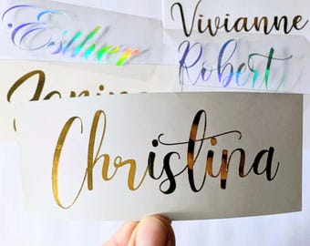 May include: Close-up of several name decals in various colors and fonts. The names include "Esther" in iridescent, "Vivianne" and "Robert" in black, and "Christina" in gold. The decals are on a white background.