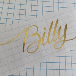 Op de afbeelding: Gouden vinyl sticker met de naam "Billy" in een cursief schrift.