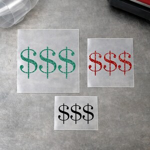 Può includere: Tre fogli quadrati di materiale per trasferimento con grafiche a forma di simbolo del dollaro. Il foglio più grande mostra tre simboli del dollaro glitterati verdi, quello medio tre glitterati rossi e il più piccolo tre simboli neri.