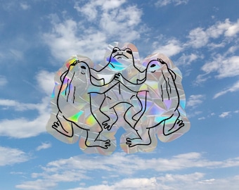 May include: A holographic sticker featuring three frogs in a circular formation, holding hands. The sticker has a reflective, iridescent surface that catches the light, displaying a black line art illustration of the frogs against a sky background.
