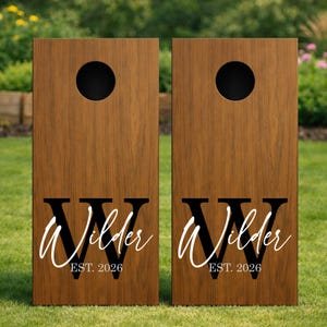 May include: Two rectangular wooden cornhole boards with a dark brown wood grain finish. Each board features a circular hole at the top and a large black letter W with the name Wilder in white script and the text EST. 2026 printed below it.