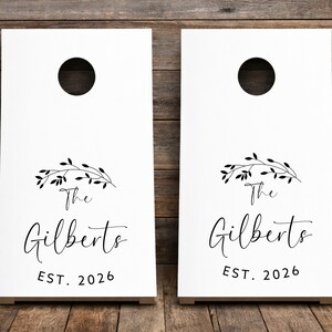Könnte beinhalten: Zwei weiße Cornhole-Boards mit einem kreisförmigen Loch im oberen Bereich. Jedes Board zeigt eine schwarze Grafik eines belaubten Zweigs über dem Text "The Gilberts" in Schreibschrift, gefolgt von dem Text "EST. 2026" in einer serifenlosen Schrift.